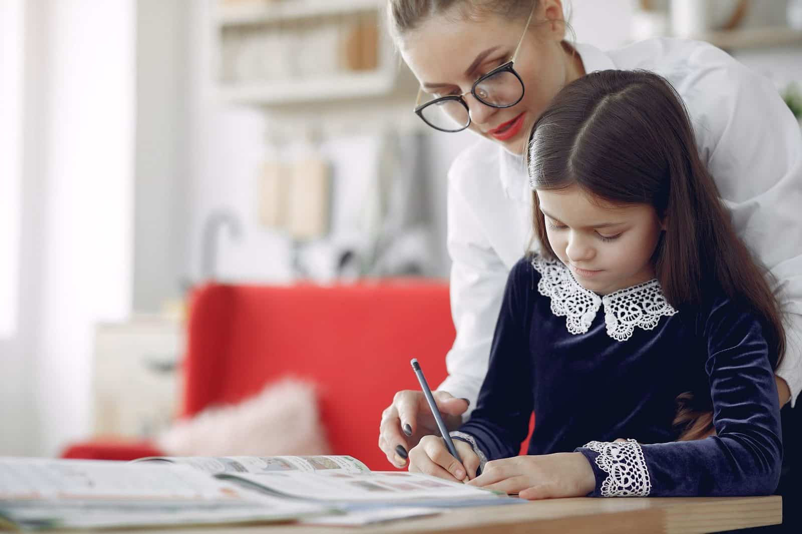 Tutor sitting beside a young student as she writes in her notebook at a bright home table