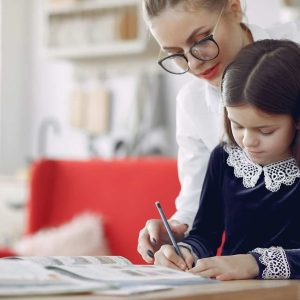 Tutor sitting beside a young student as she writes in her notebook at a bright home table